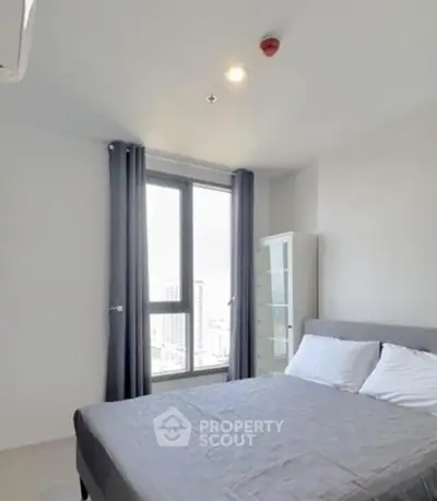 Modern bedroom with large window offering city views, featuring a cozy bed and minimalist decor.