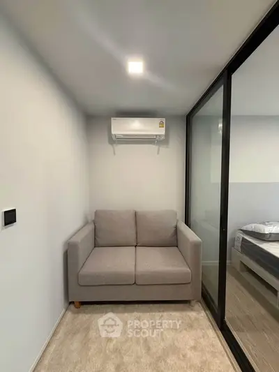 Modern compact living room with gray sofa and air conditioning, adjacent to a bedroom with glass partition.