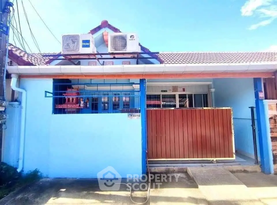 Charming single-story home with blue facade and red roof, featuring air conditioning units and a gated entrance.