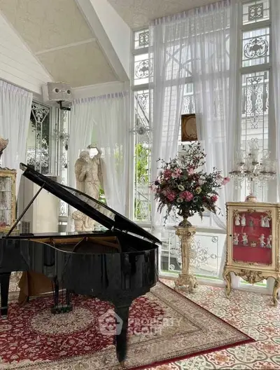 Elegant living room with grand piano and vintage decor, featuring ornate windows and floral arrangements.