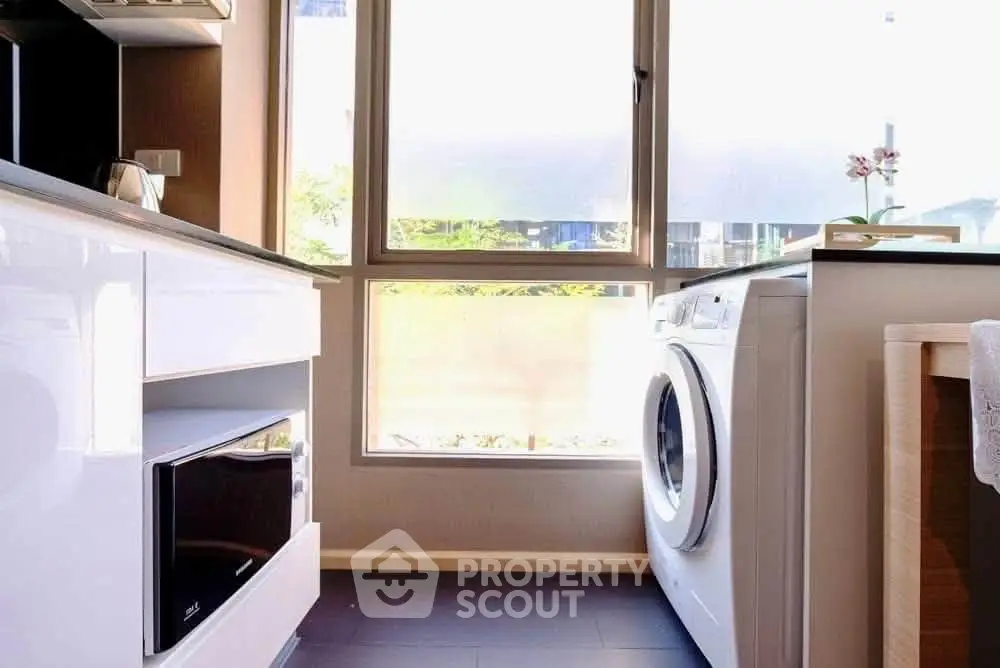 Modern kitchen with washing machine and microwave, bright natural light through large window.