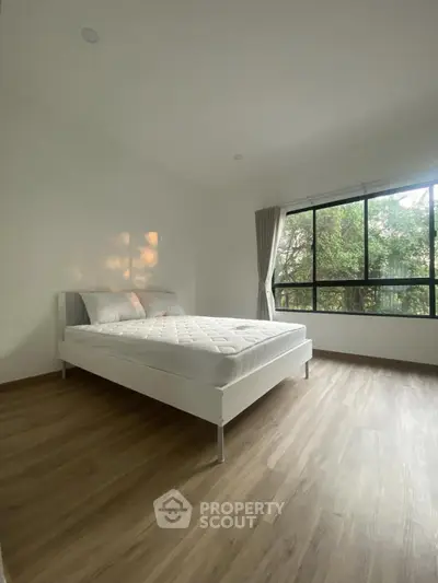 Spacious bedroom with large window and natural light, featuring a minimalist design and wooden flooring.