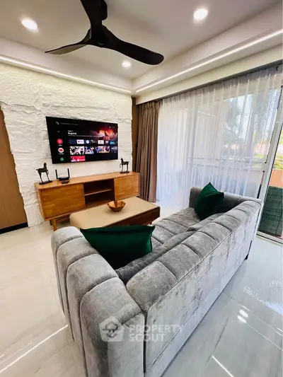 Modern living room with plush gray sofa, wall-mounted TV, and sliding glass doors leading to a balcony.