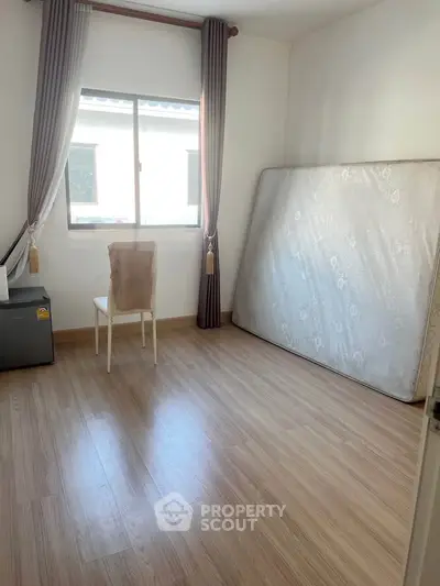Spacious bedroom with wooden flooring and natural light, featuring a window view and minimal furnishings.