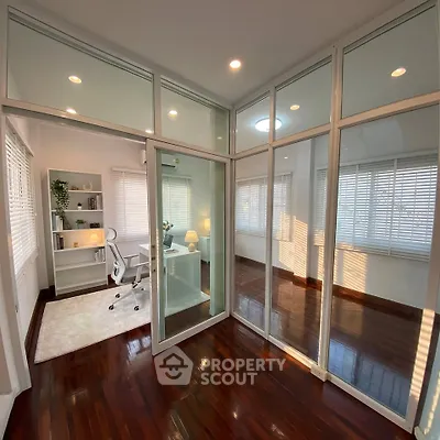 Modern home office with glass walls and wooden flooring, featuring a sleek white desk and ergonomic chair.