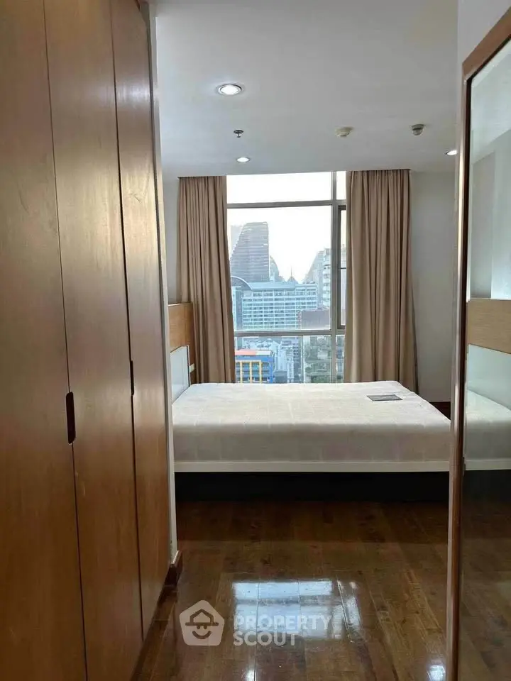 Modern bedroom with city view, wooden flooring, and large window in urban apartment.
