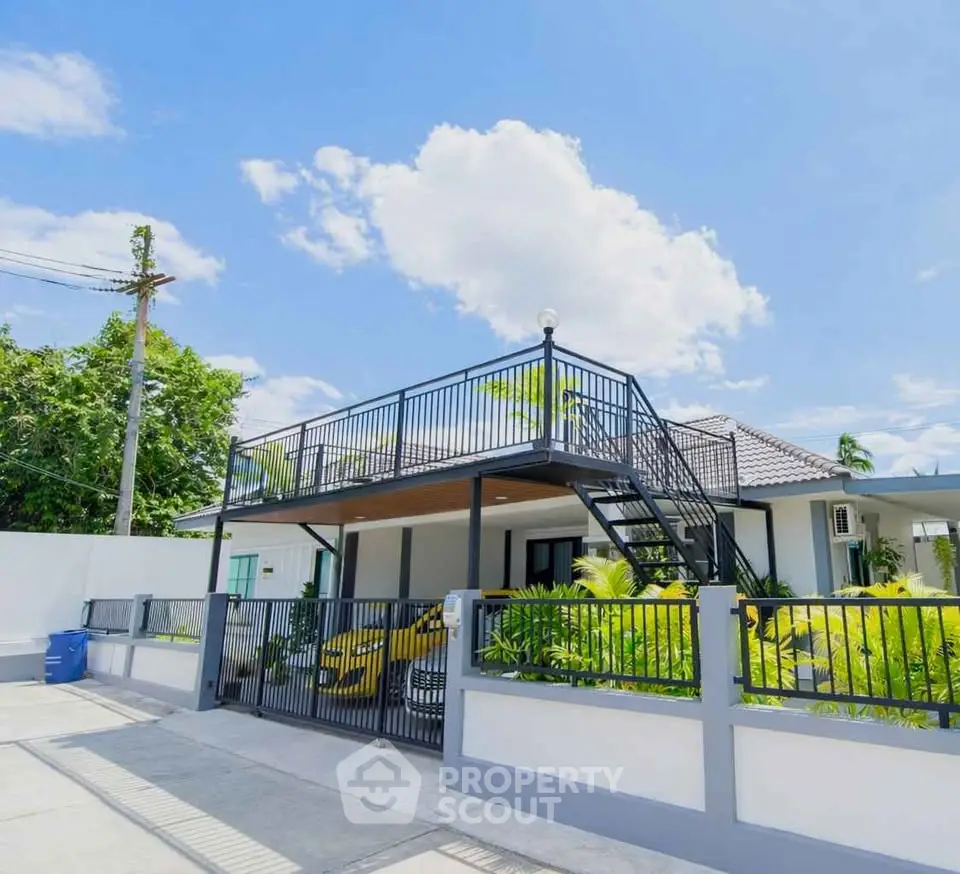 Modern two-story house with rooftop terrace and lush garden under clear blue sky.