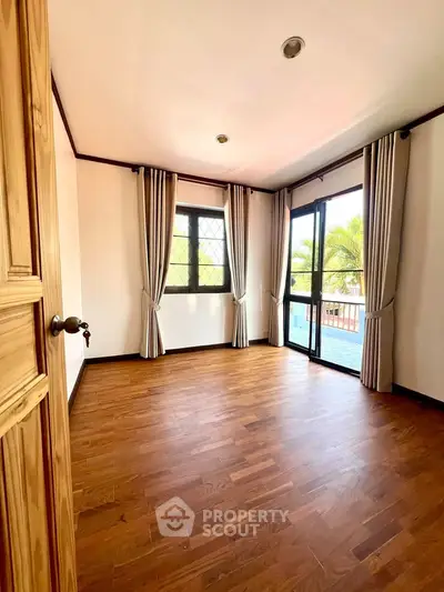 Bright empty room with wooden floors and large windows overlooking greenery.