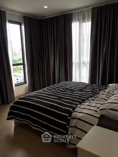 Modern bedroom with large windows and city view, featuring stylish striped bedding and dark curtains.