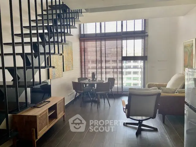 Modern living room with high ceiling and stylish staircase, featuring large windows and chic furniture.