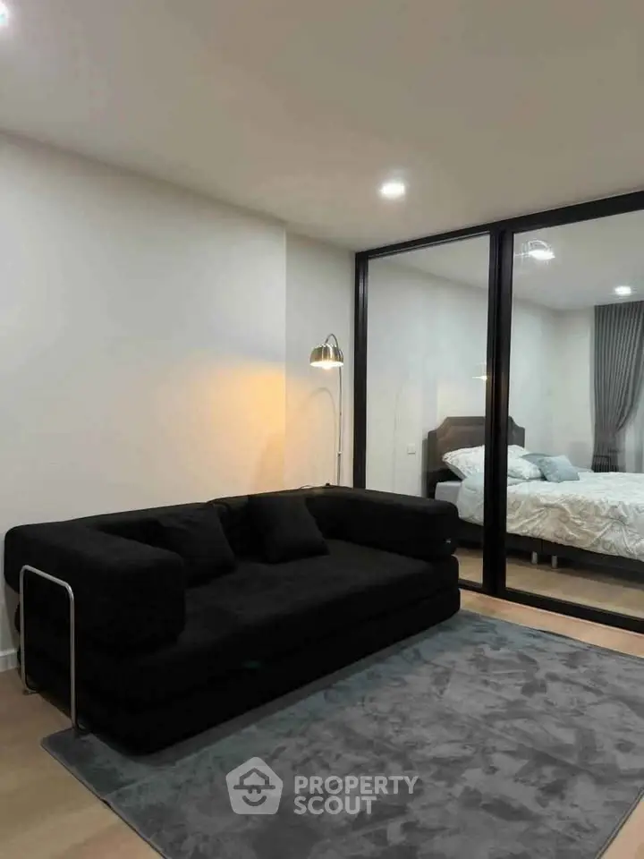 Modern living room with sleek black sofa and view into cozy bedroom through glass partition.