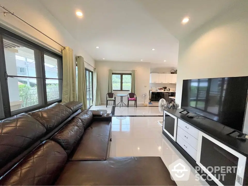 Spacious living room with modern leather sofa and large TV, open to kitchen area with natural light.