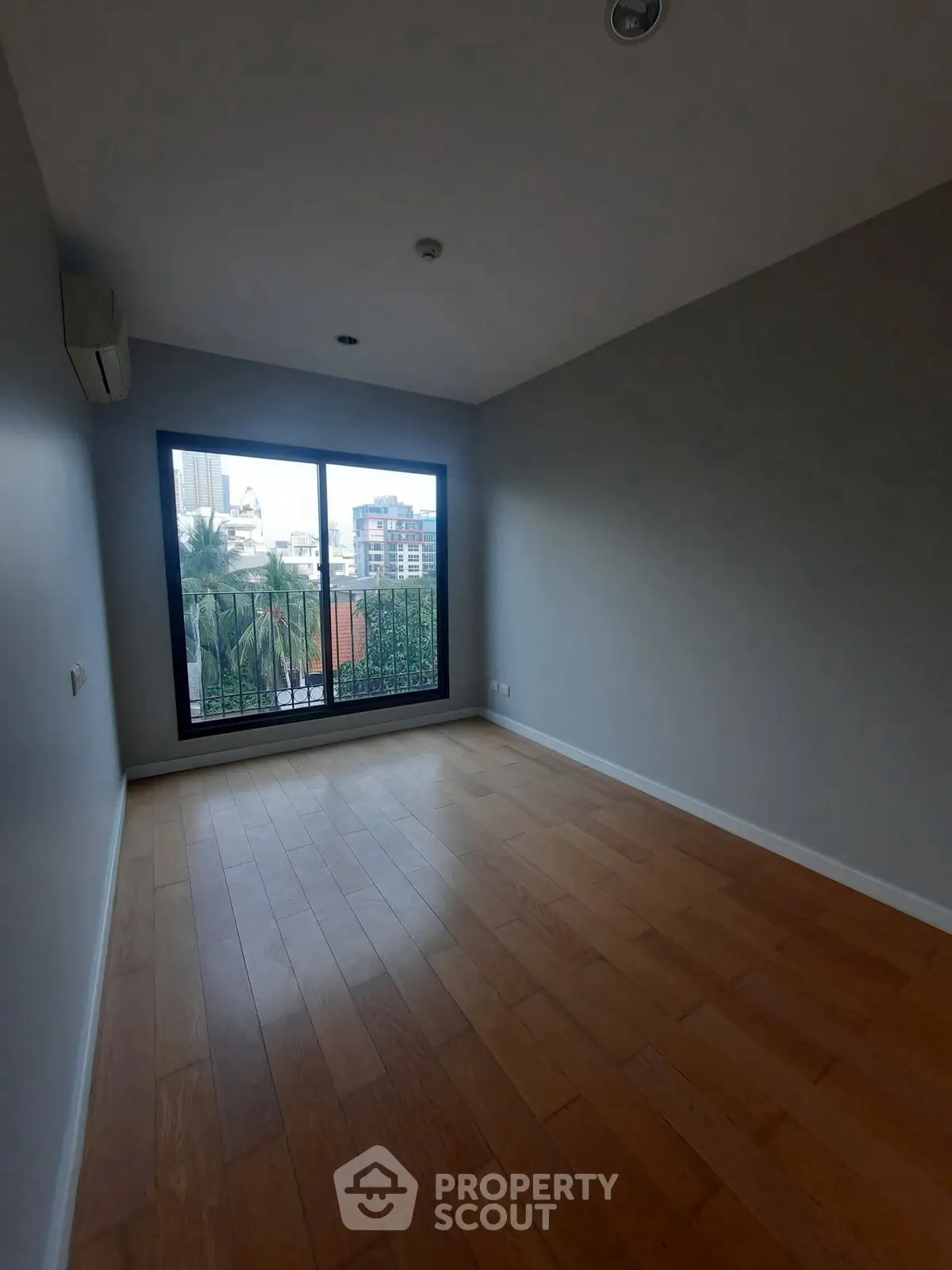 Spacious empty room with wooden flooring and large window offering city view.