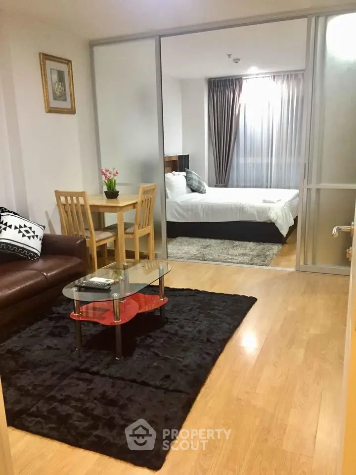 Cozy living room with wooden flooring and glass coffee table leading to a stylish bedroom with plush bedding.