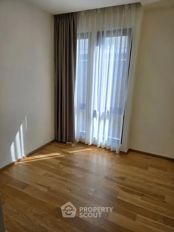 Bright empty room with wooden flooring and large window with curtains.