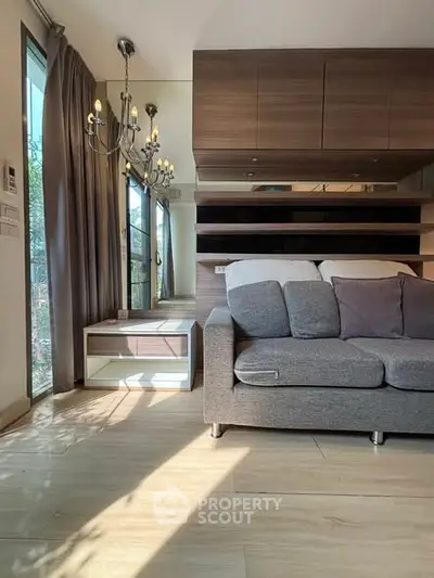 Modern living room with elegant chandelier and cozy gray sofa, featuring wooden flooring and large windows.