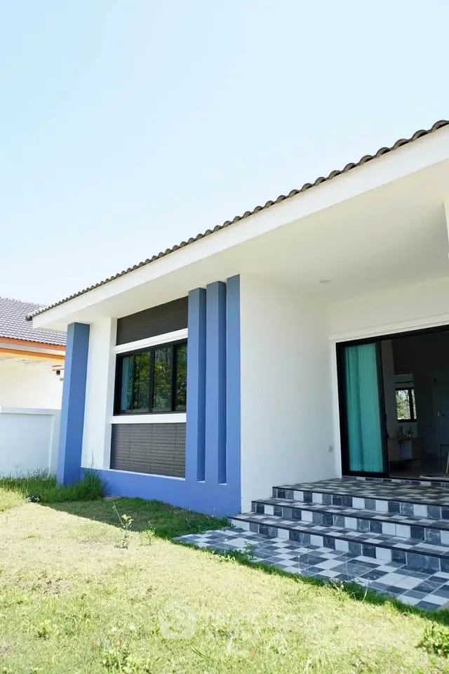 Modern single-story house with blue accents and tiled steps in sunny garden setting.