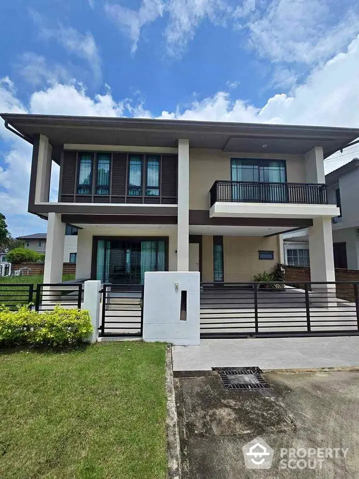 Modern two-story house with large windows and balcony, surrounded by lush greenery and a spacious driveway.