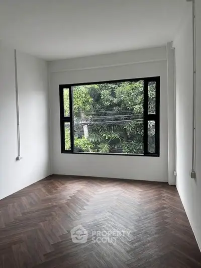 Spacious room with large window and herringbone wood flooring, offering lush green views.