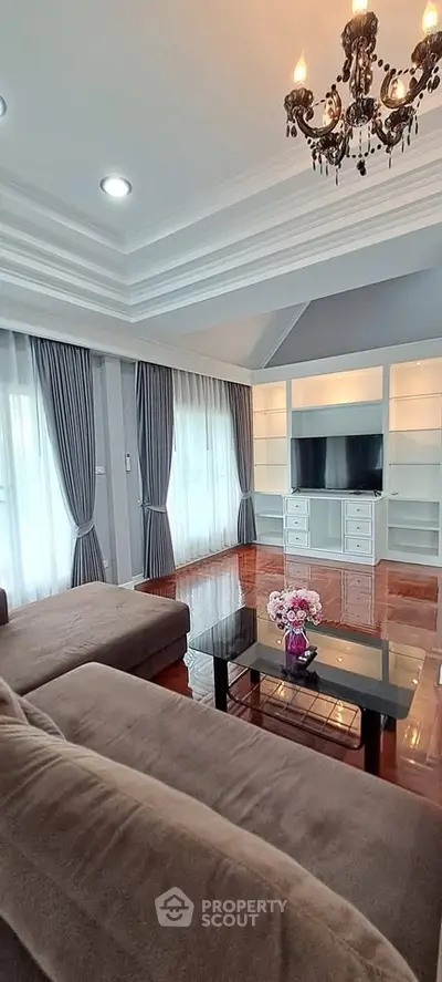 Elegant living room with chandelier, large sofa, and built-in TV unit, featuring floor-to-ceiling windows and stylish curtains.