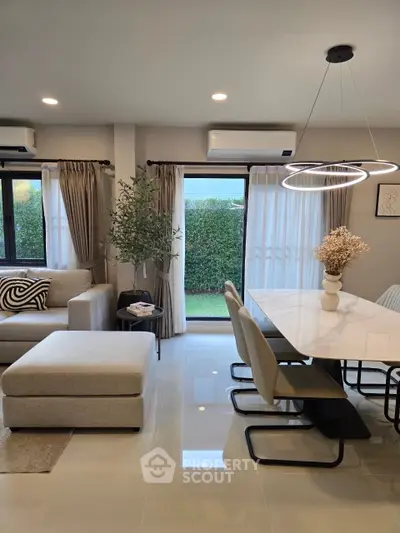 Modern living room with elegant dining area and stylish lighting, featuring large windows and lush garden view.