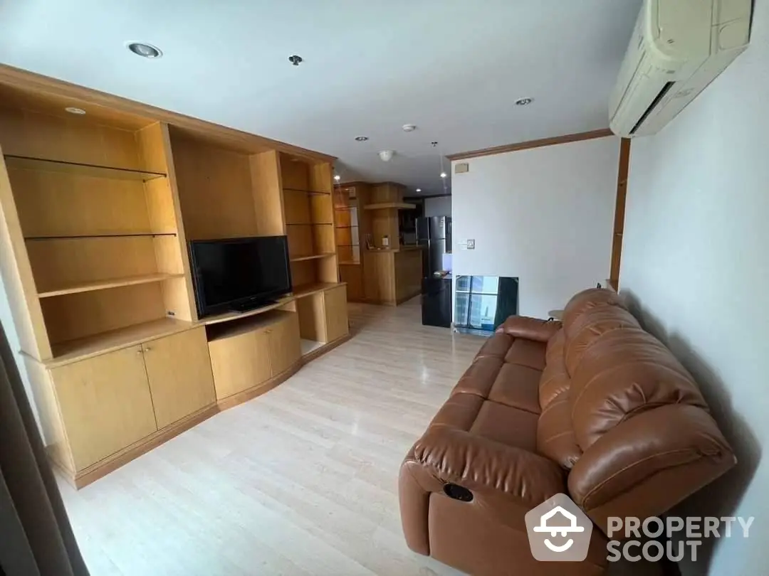 Spacious living room with large leather sofa and built-in wooden entertainment center, leading to an open kitchen area.