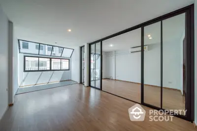 Spacious and modern living room with large windows and reflective glass doors leading to an adjoining room, featuring sleek wooden flooring and ample natural light.