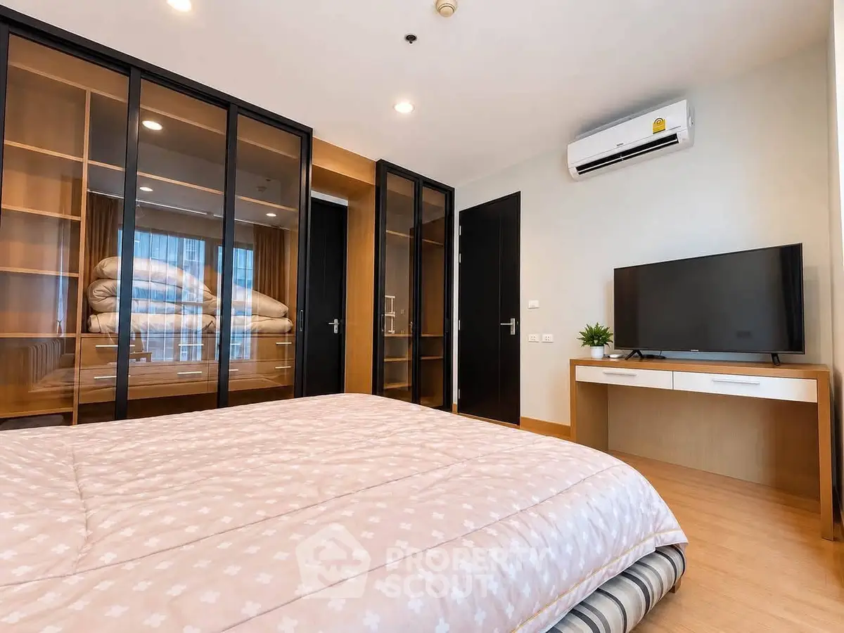 Modern bedroom with glass wardrobe and TV, featuring cozy bedding and air conditioning.
