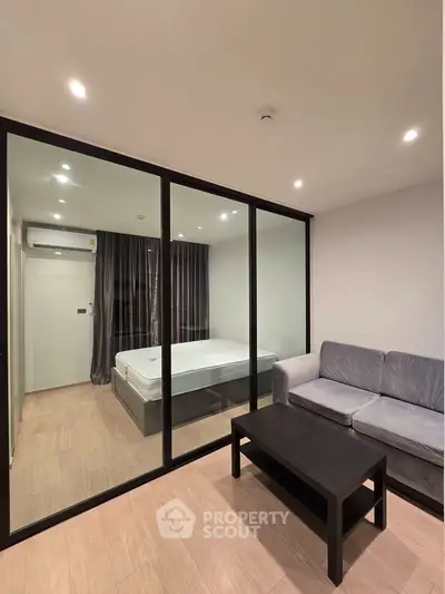 Modern living room with glass partition and cozy bedroom view, featuring sleek furniture and ambient lighting.