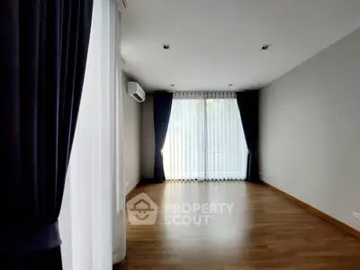 Spacious empty room with wooden flooring and large windows with curtains.