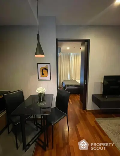 Elegant living space with modern dining set, sleek pendant lighting, and warm wooden flooring extending into a cozy bedroom with soft lighting.