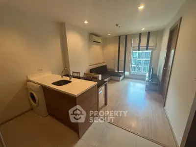 Modern open-layout living room with kitchen and washing machine, featuring wooden flooring and natural light.