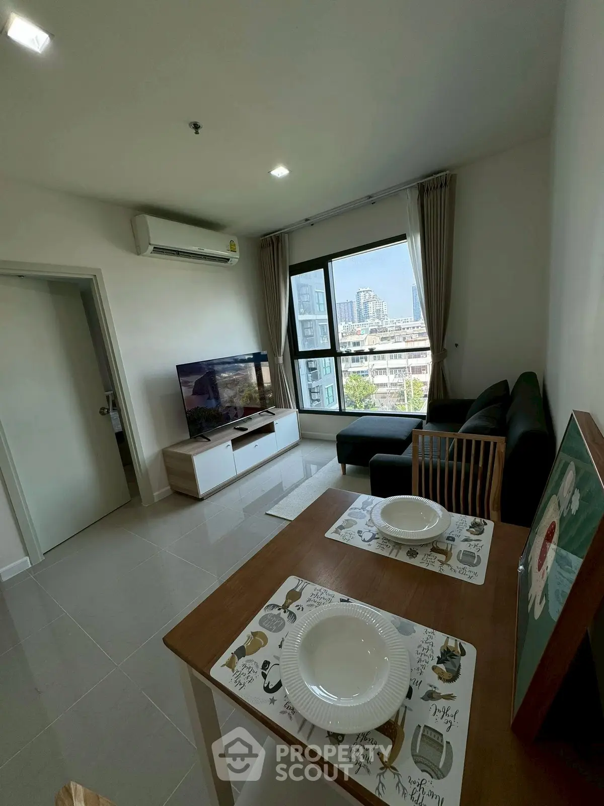 Modern living room with city view, featuring sleek furniture and dining area in a bright apartment.
