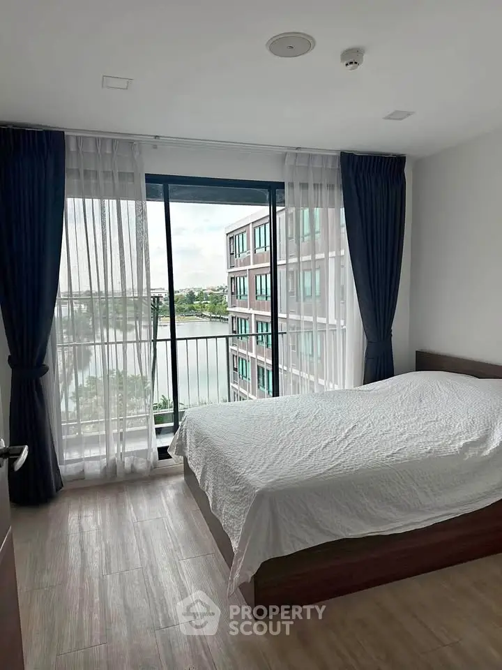 Spacious bedroom with large window offering stunning view of modern building and serene water.