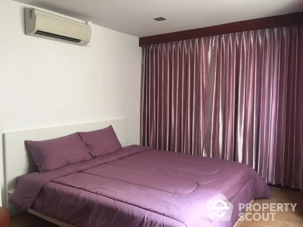 Cozy bedroom with purple bedding and curtains, featuring air conditioning and wooden flooring.