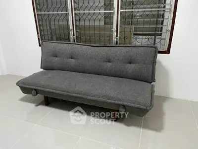Modern gray sofa in a minimalist living room with tiled flooring and barred windows.