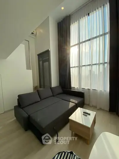 Modern living room with high ceilings and large windows, featuring a sleek gray sofa and natural light.