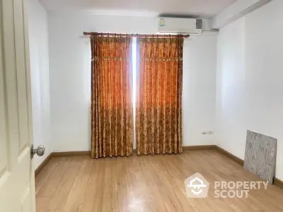 Spacious and well-lit empty room with polished wooden flooring, large windows draped with orange curtains, and a clean white ceiling with an air conditioning unit.