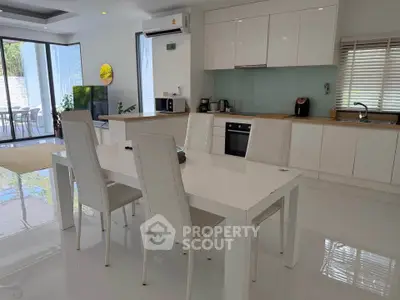 Modern open-layout kitchen with sleek white furniture and appliances, featuring a dining area and large windows.