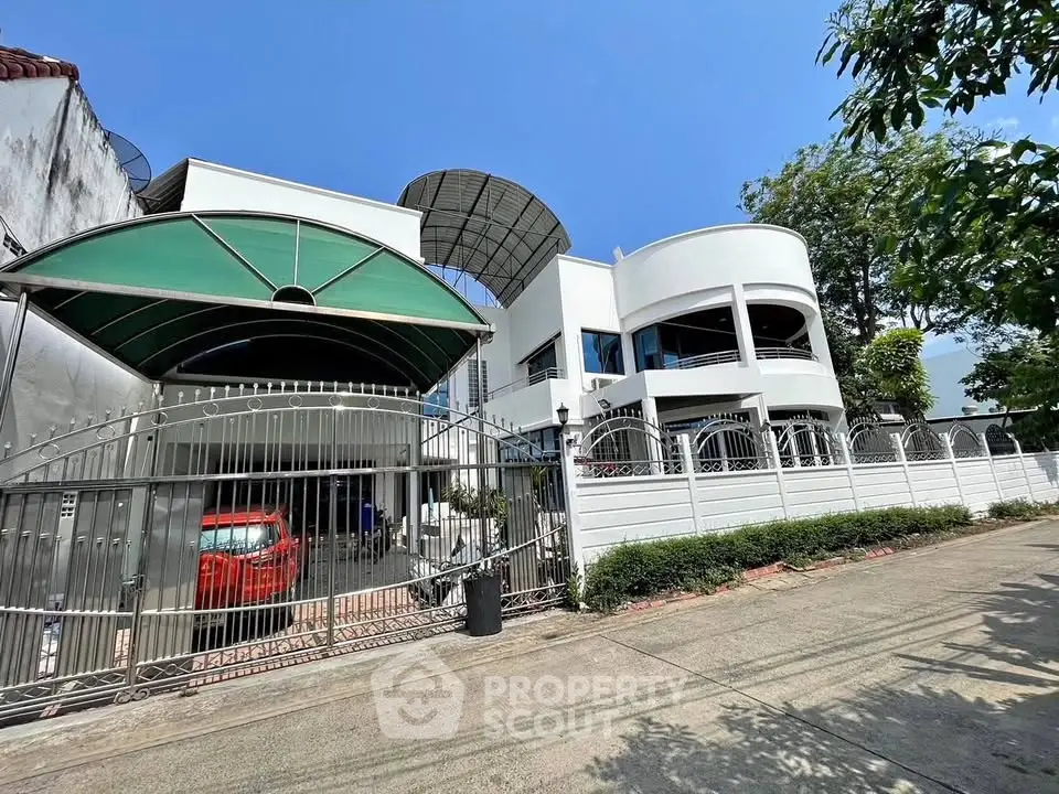 Modern two-story house with gated entrance and carport, featuring unique architectural design and lush greenery.
