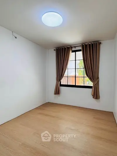 Spacious empty room with wooden flooring and large window with curtains.