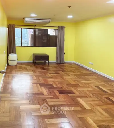Spacious living room with parquet flooring and vibrant yellow walls, featuring a large window with city view.