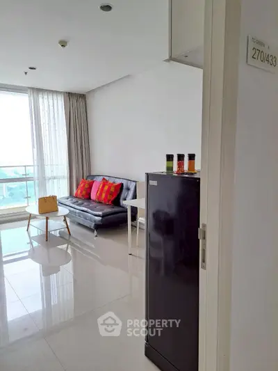 Modern living room with sleek sofa and vibrant cushions, featuring a compact fridge and large window for natural light.