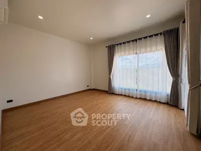 Spacious empty room with wooden flooring and large window with curtains.
