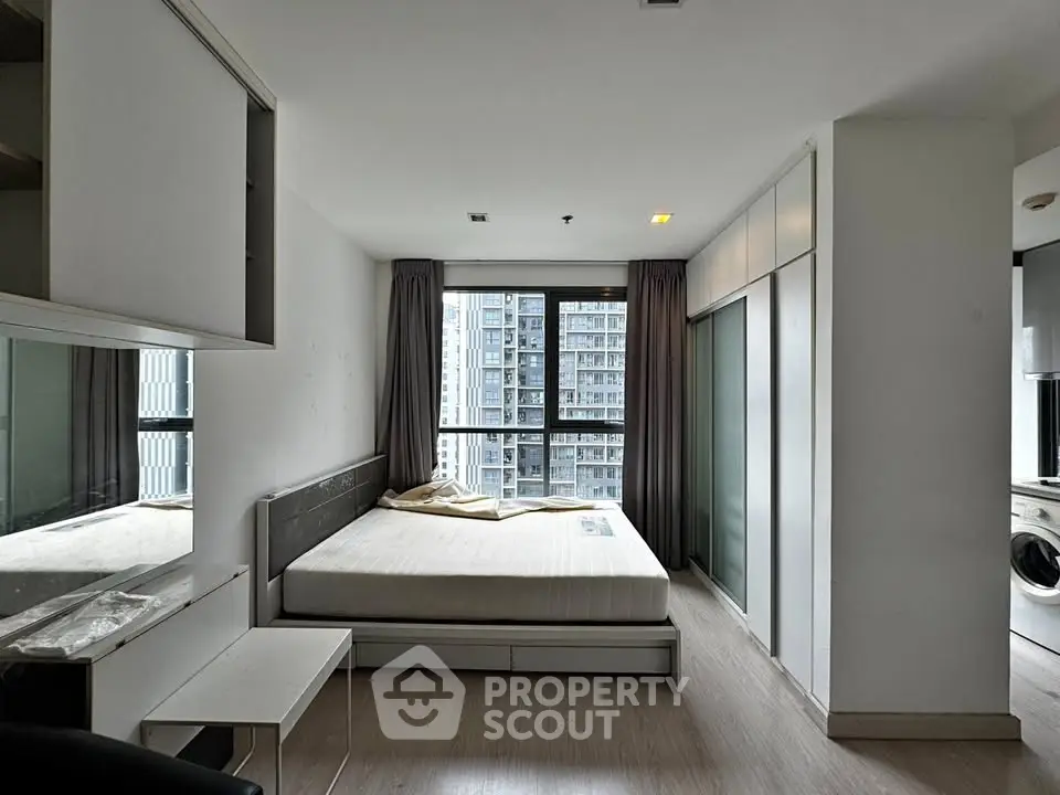 Modern bedroom with large window and city view, featuring sleek furniture and built-in storage.