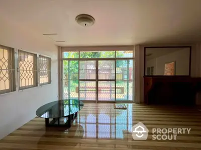 Spacious living room with polished wooden floors, large windows offering ample natural light, and a sleek glass coffee table, perfect for modern living.