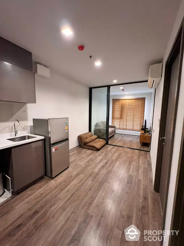 Spacious modern studio apartment with sleek kitchen, full-sized fridge, and cozy sitting area leading to a sunlit balcony with wooden accents.