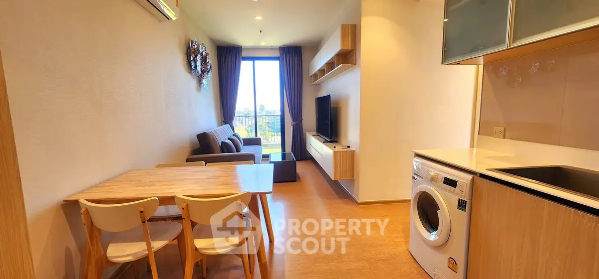 Modern open-plan living room with kitchen and washing machine, featuring a cozy sofa and large window view.