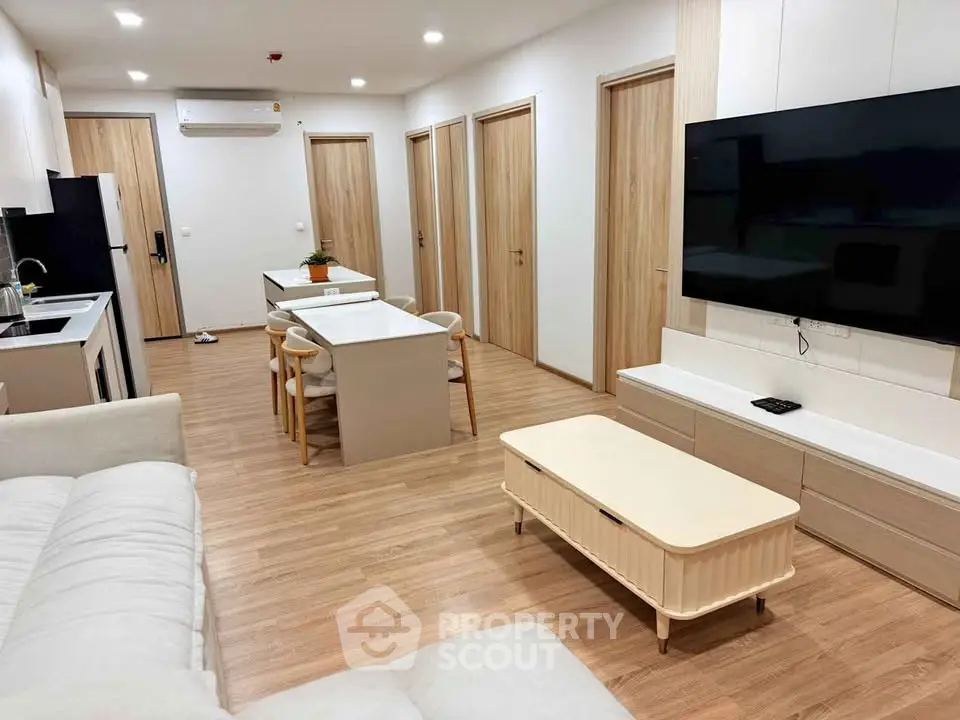 Modern open-layout living room with sleek kitchen and dining area, featuring contemporary furniture and large TV.