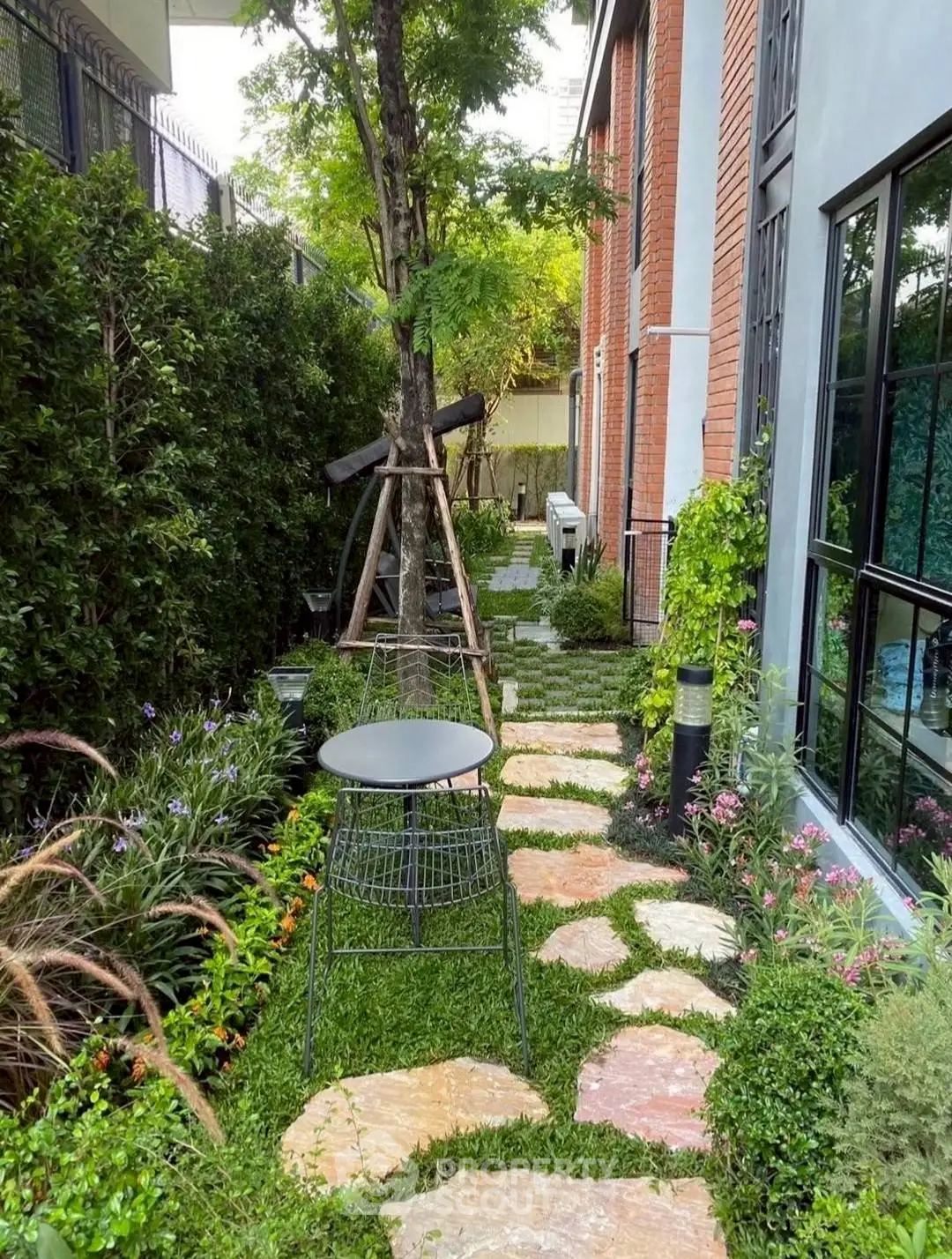 Charming garden pathway beside modern brick building with lush greenery and outdoor seating.